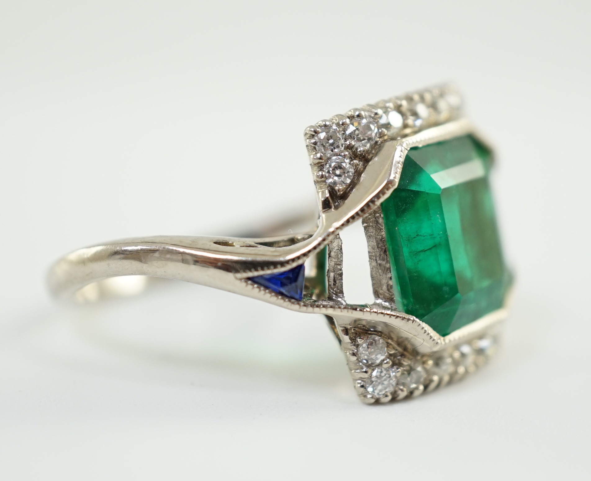 An Art Deco 18k white gold, emerald and diamond set square cluster dress ring, with shaped cut sapphire set shoulders, (a.f.)